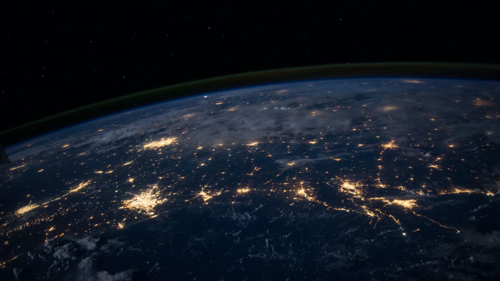 Vista nocturna de la Tierra desde el espacio, mostrando ciudades y continentes iluminados. La curvatura de la Tierra es visible, con un cielo oscuro y estrellas dispersas en el cielo. Una delgada línea verde de resplandor atmosférico recorre el horizonte.