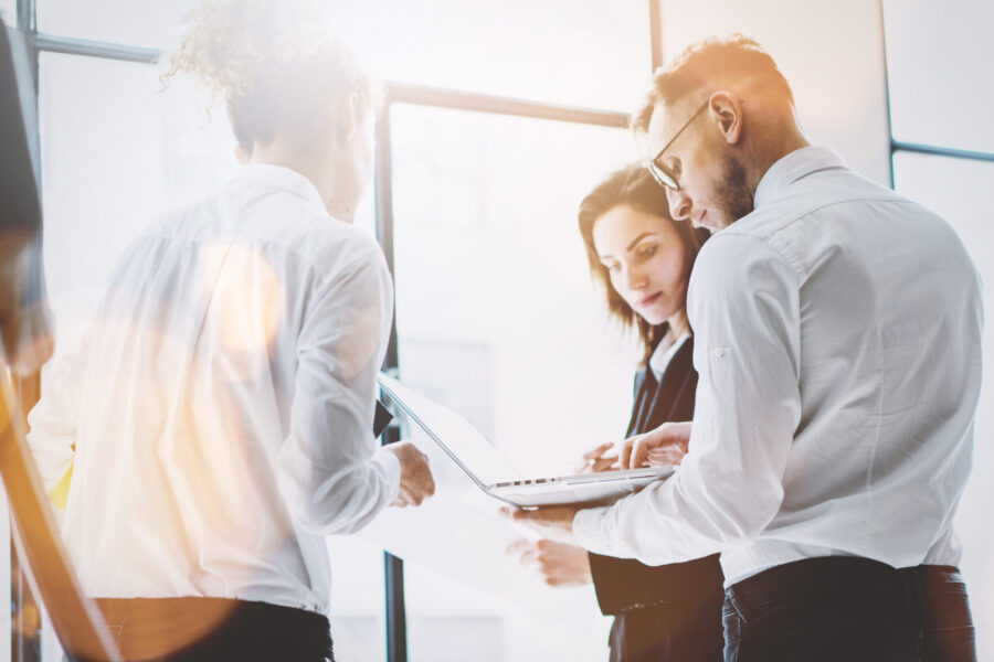 Tres personas vestidas de negocios conversan junto a grandes ventanales. Una sostiene una computadora portátil, mientras que otra parece estar revisando documentos. La luz del sol se cuela por las ventanas, proyectando un resplandor brillante sobre la escena.