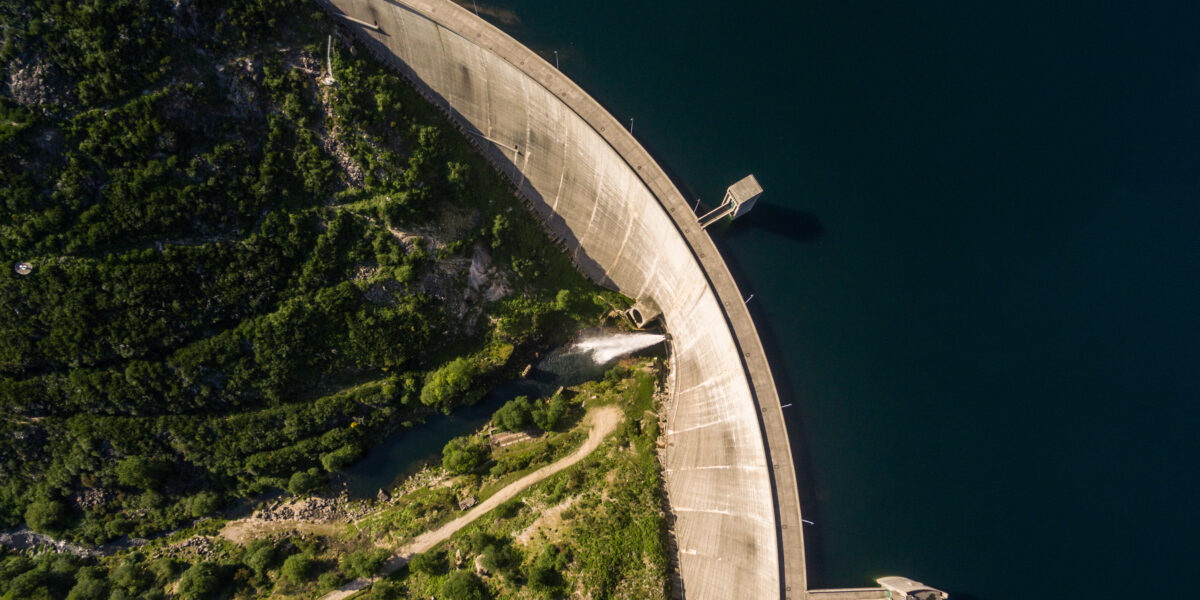 Vista aérea de una gran presa que se curva sobre un cuerpo de agua de color azul intenso. Una exuberante vegetación rodea la estructura, con un pequeño camino y un río visibles a la izquierda. El muro de la presa proyecta una sombra sobre el agua.