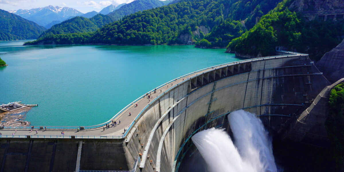Una gran presa de hormigón vierte agua a un río que se encuentra abajo, rodeado de exuberantes montañas verdes. Un embalse turquesa se divisa sobre la presa bajo un cielo parcialmente nublado. Se pueden ver pequeñas figuras de personas caminando por la cima de la presa.