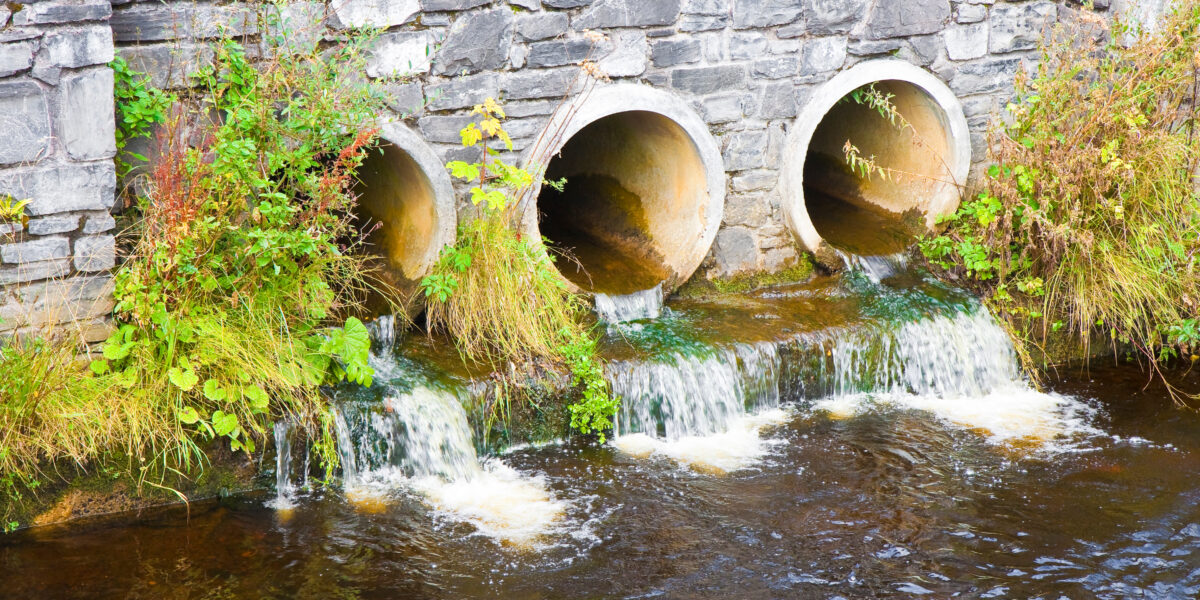 Tres tuberías de hormigón empotradas en un muro de piedra vierten agua cristalina a un río. La zona alrededor de las tuberías luce un follaje verde. El agua fluyente forma pequeñas cascadas al desembocar en el río.