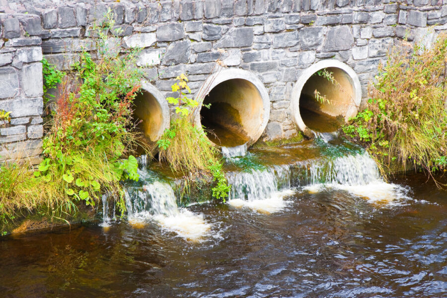 Tres tuberías de hormigón empotradas en un muro de piedra vierten agua cristalina a un río. La zona alrededor de las tuberías luce un follaje verde. El agua fluyente forma pequeñas cascadas al desembocar en el río.