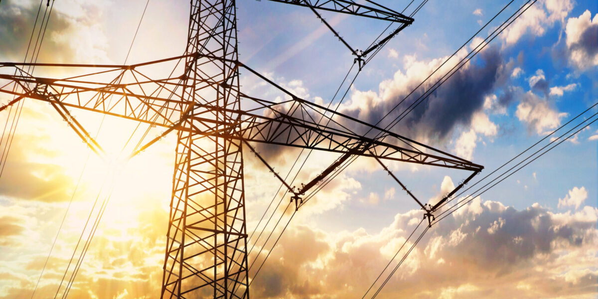 Una alta torre de electricidad metálica se alza contra un cielo vibrante, con el sol brillando a través de su estructura. Las líneas eléctricas se extienden hacia afuera, y el cielo presenta nubes con una mezcla de tonos azules y naranjas, indicando el amanecer o el atardecer.