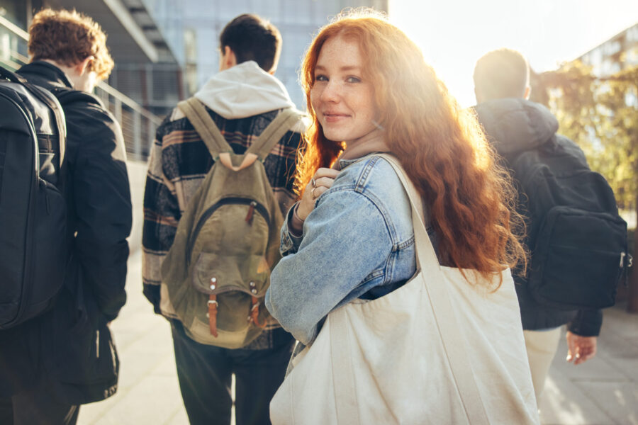 Una joven pelirroja de pelo largo, vestida con una chaqueta vaquera, sonríe mientras camina con tres personas. Lleva una bolsa de mano al hombro. Están al aire libre, con edificios modernos al fondo y la luz del sol filtrándose.