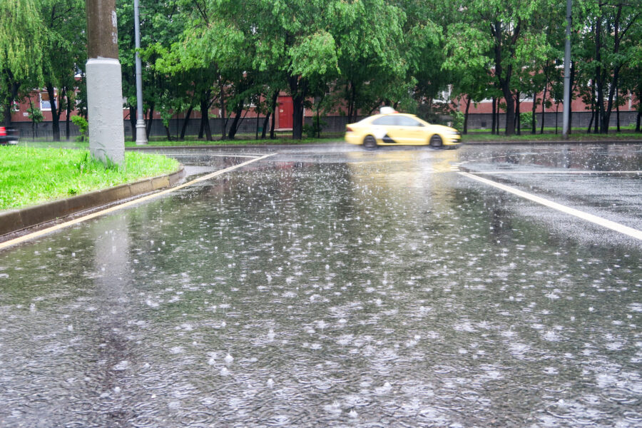 Un taxi amarillo avanza a toda velocidad por una calle lluviosa y casi vacía. Las gotas de lluvia ondulan sobre los grandes charcos de la carretera. Árboles verdes y un edificio rojo al fondo contrastan con la escena.
