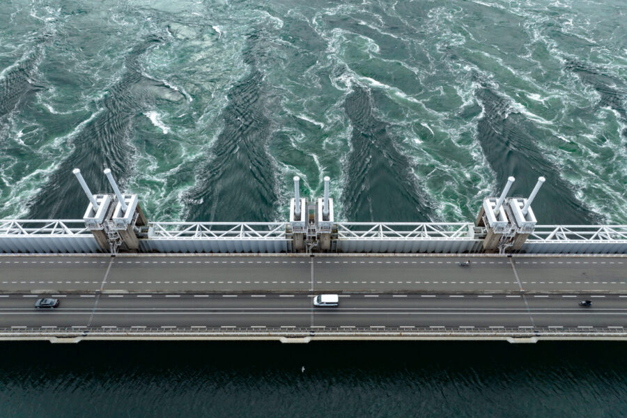 Vista aérea de un puente que cruza una masa de agua con remolinos. El puente tiene vehículos circulando y presenta varios elementos estructurales verticales. El agua debajo muestra patrones de corrientes u olas.