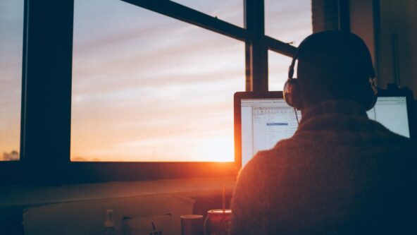 Una persona con auriculares está sentada en un escritorio trabajando en una computadora, mirando a un gran ventanal. La puesta de sol proyecta una cálida luz en la habitación, creando un ambiente acogedor. Varios objetos están colocados sobre el escritorio, frente a ella.