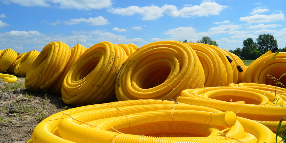 Grandes rollos de tubos de plástico amarillo se apilan en un campo abierto bajo un cielo azul brillante con nubes dispersas. Los tubos están dispuestos en filas ordenadas, creando un patrón repetitivo contra el fondo natural de árboles a lo lejos.