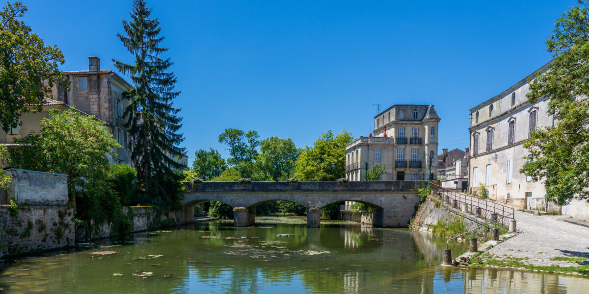 Vista de un río tranquilo con un puente de piedra arqueado, rodeado de edificios antiguos y árboles en un día soleado. El cielo es azul claro y el reflejo de la escena se refleja en el agua.