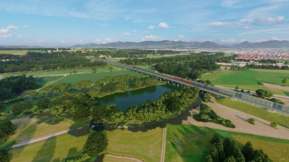 Vista aérea de un paisaje con un largo puente sobre un río, rodeado de exuberantes campos verdes y árboles. Al fondo se ven colinas ondulantes bajo un cielo azul parcialmente nublado.