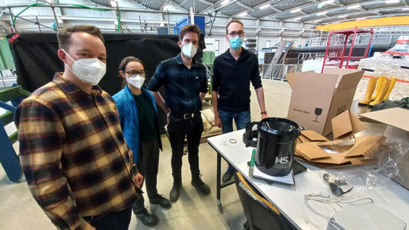 Cuatro personas con mascarillas están de pie alrededor de una mesa en un amplio espacio interior con un techo alto y curvo. Sobre la mesa hay un objeto cilíndrico negro y varias cajas de cartón abiertas. Parecen estar en un taller o laboratorio.