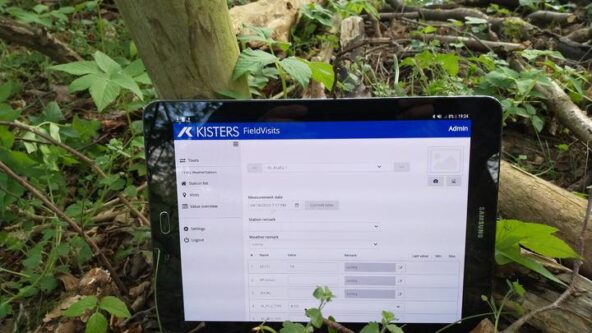 Una tableta muestra una aplicación de visita de campo con menús y entradas de datos visibles. Está apoyada contra el tronco de un árbol, rodeada de plantas verdes y ramas caídas en un entorno forestal.