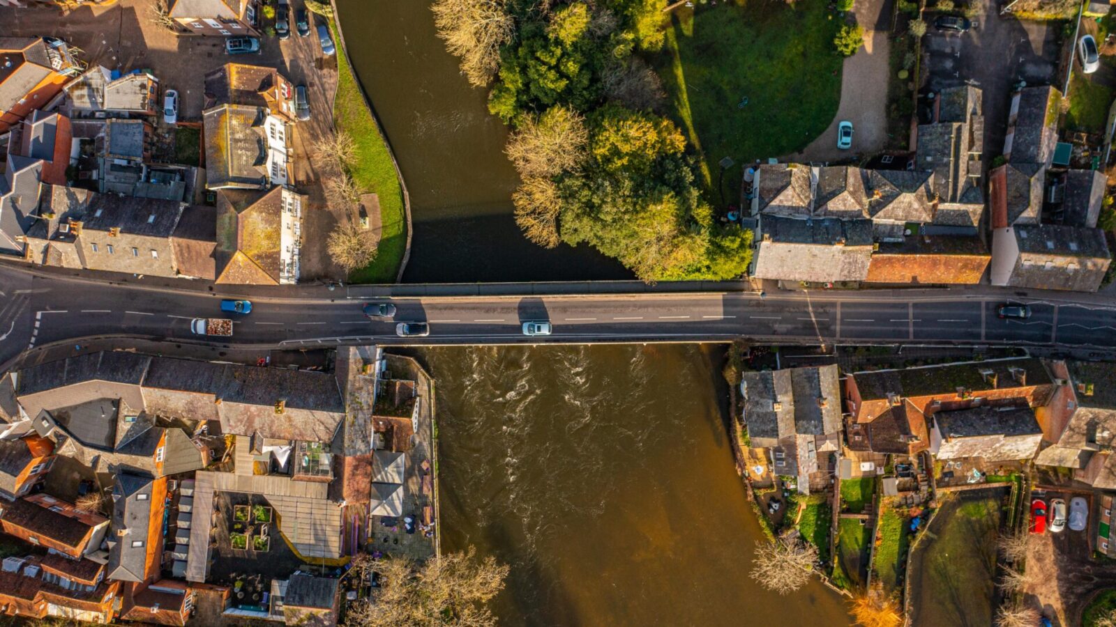 Vista aérea de un pequeño pueblo atravesado por un río. Un puente vial cruza el río, rodeado de edificios y vegetación. Se ven coches en el puente y las calles, con árboles a la orilla.