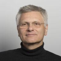 Una persona con cabello gris y gafas viste un cuello alto negro y sonríe levemente sobre un fondo gris claro.