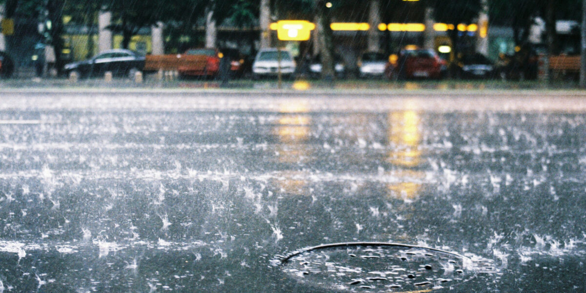 Las gotas de lluvia salpican una calle mojada, con una tapa de alcantarilla visible en primer plano. Al fondo, borroso, se ven coches aparcados y letreros iluminados de tiendas bajo una copa de árboles.