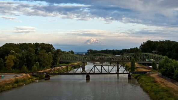 Una vista panorámica de un río con dos puentes metálicos verdes que lo cruzan. A lo lejos, se divisa una montaña nevada bajo un cielo parcialmente nublado. Una exuberante vegetación bordea las orillas del río.