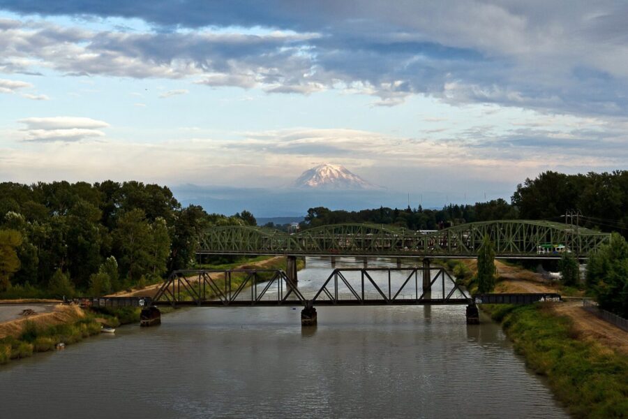 Una vista panorámica de un río con dos puentes metálicos verdes que lo cruzan. A lo lejos, se divisa una montaña nevada bajo un cielo parcialmente nublado. Una exuberante vegetación bordea las orillas del río.