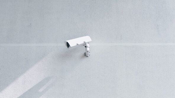 Una cámara de seguridad blanca montada en una pared minimalista de hormigón gris claro. La cámara proyecta una sombra a la izquierda, realzando la textura y la simplicidad de la pared.