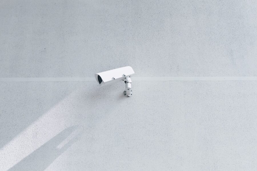 Una cámara de seguridad blanca montada en una pared minimalista de hormigón gris claro. La cámara proyecta una sombra a la izquierda, realzando la textura y la simplicidad de la pared.