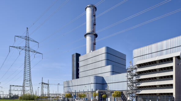 Una gran central eléctrica industrial con una alta chimenea cilíndrica se muestra contra un cielo azul despejado. Se ven líneas eléctricas de alta tensión y estructuras metálicas, con zonas de césped y pequeños árboles en primer plano.