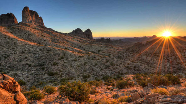 Atardecer sobre un paisaje desértico con montañas escarpadas y vegetación escasa. El sol proyecta un cálido resplandor y largas sombras que realzan la textura del terreno.