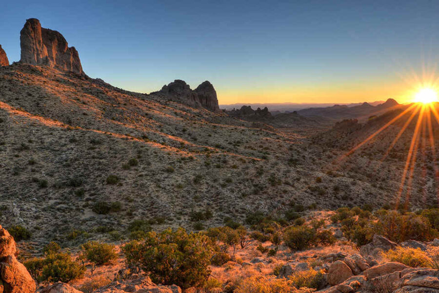 Atardecer sobre un paisaje desértico con montañas escarpadas y vegetación escasa. El sol proyecta un cálido resplandor y largas sombras que realzan la textura del terreno.