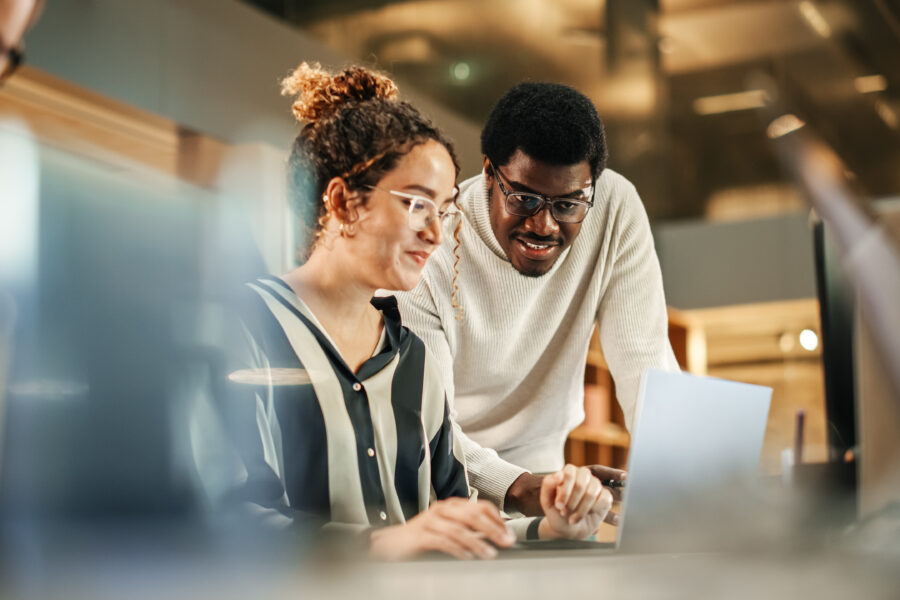 Dos personas trabajan juntas en un escritorio, concentradas en la pantalla de una laptop. La persona de la izquierda está sentada, con gafas y una camisa a rayas. La persona de la derecha está de pie, sonriendo, con un suéter blanco. El entorno parece ser una oficina.