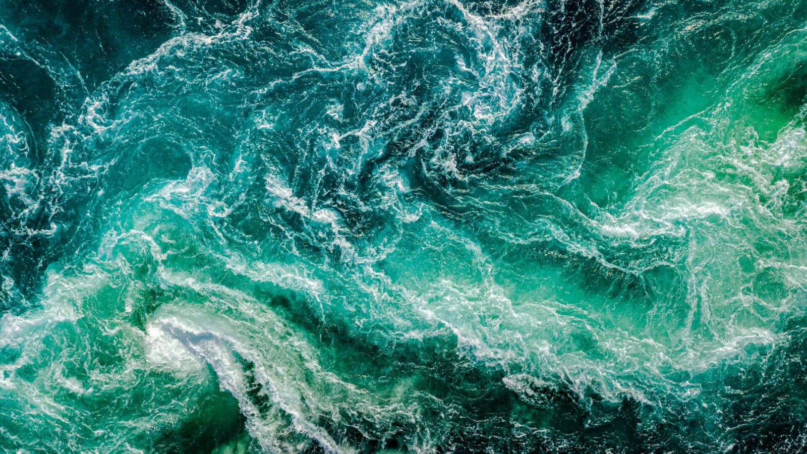 Vista aérea de las olas turquesas del océano, que se arremolinan creando patrones intrincados. El movimiento y la mezcla de la espuma blanca entre los tonos azul oscuro y claro transmiten una sensación de energía dinámica y belleza natural.