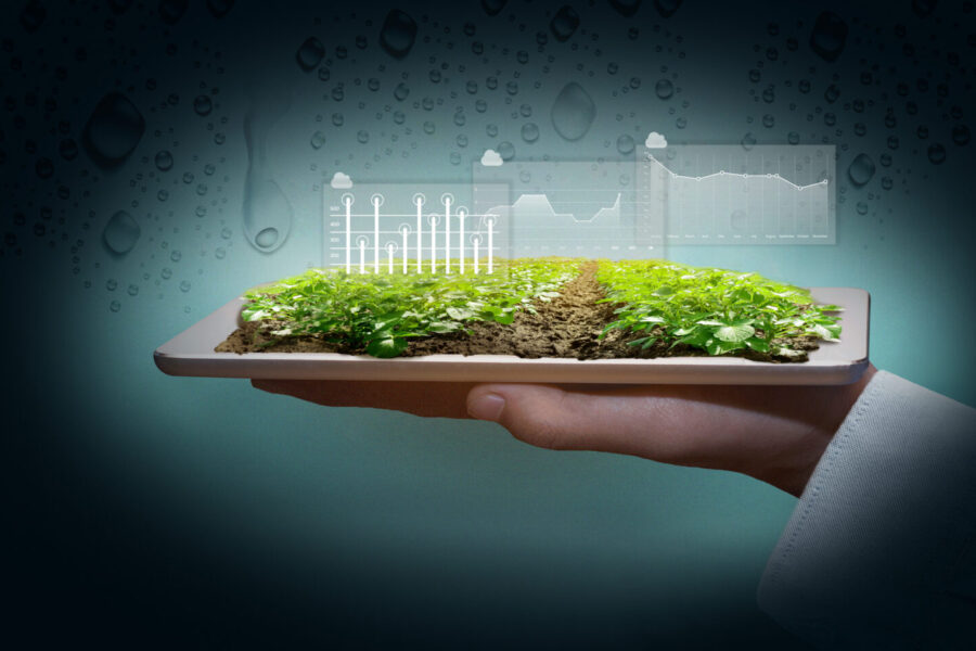 Una mano sostiene una tableta que muestra un campo verde en miniatura con pequeñas plantas que crecen en la tierra. Los gráficos superpuestos muestran tablas y gráficos de datos que indican análisis agrícolas o ambientales. El fondo es oscuro con patrones de gotas de agua.