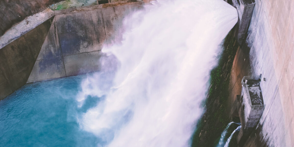 El agua brota con fuerza desde la salida de una gran presa hacia una poza inferior, mostrando su potente caudal. La estructura de hormigón está parcialmente cubierta de musgo, y el agua parece espumosa y turbulenta al caer en cascada.