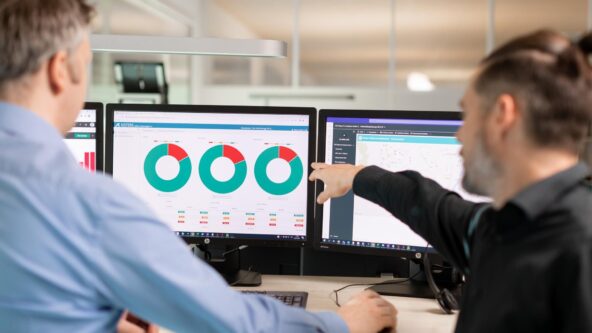 Dos personas en una oficina analizan datos en dos monitores. La pantalla izquierda muestra tres gráficos circulares con segmentos verdes, rojos y azules. La pantalla derecha muestra un mapa. Una persona señala el monitor derecho y comenta los datos.