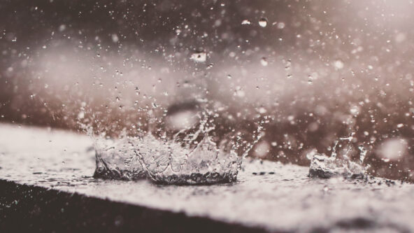 Primer plano de gotas de lluvia salpicando una superficie, creando una especie de corona. La imagen captura el movimiento dinámico y las gotas suspendidas en el aire, con un fondo difuminado en tonos suaves y neutros.