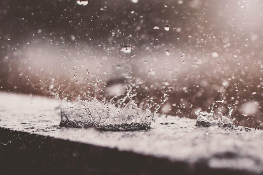 Primer plano de gotas de lluvia salpicando una superficie, creando una especie de corona. La imagen captura el movimiento dinámico y las gotas suspendidas en el aire, con un fondo difuminado en tonos suaves y neutros.