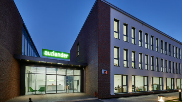 Moderno edificio de oficinas al anochecer, con amplios ventanales y fachada de ladrillo y blanco. La entrada tiene una fachada de cristal con el nombre