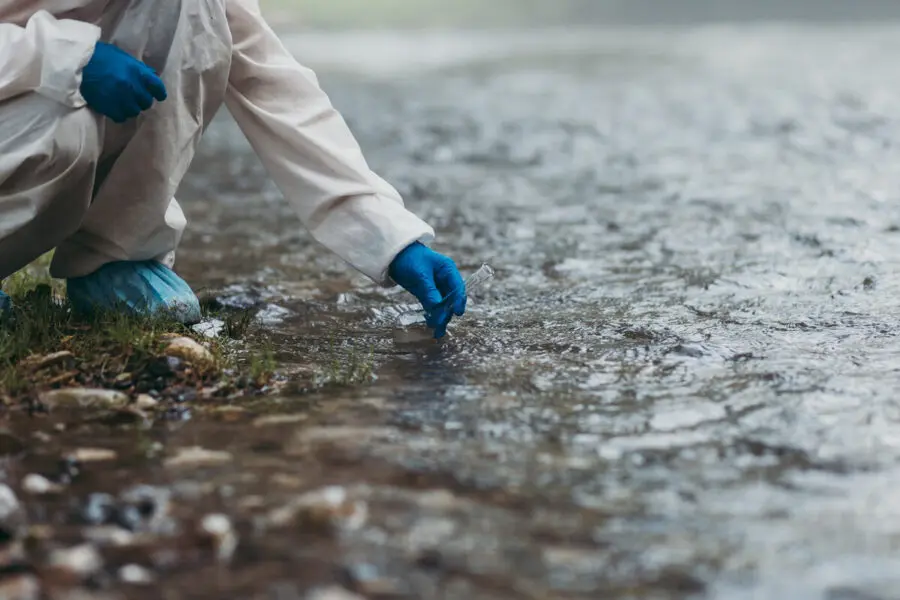 Una persona que viste un traje protector blanco, guantes azules y cubrezapatos se agacha al borde de un cuerpo de agua y extiende la mano para tocar o recoger una muestra de la superficie clara y fluida.