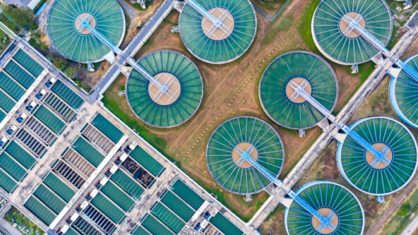 Vista aérea de una planta de tratamiento de aguas residuales con clarificadores circulares y tanques de sedimentación rectangulares. Las estructuras circulares verdes se distribuyen uniformemente en una cuadrícula, interconectadas por pasarelas, rodeadas de césped y equipos adicionales.