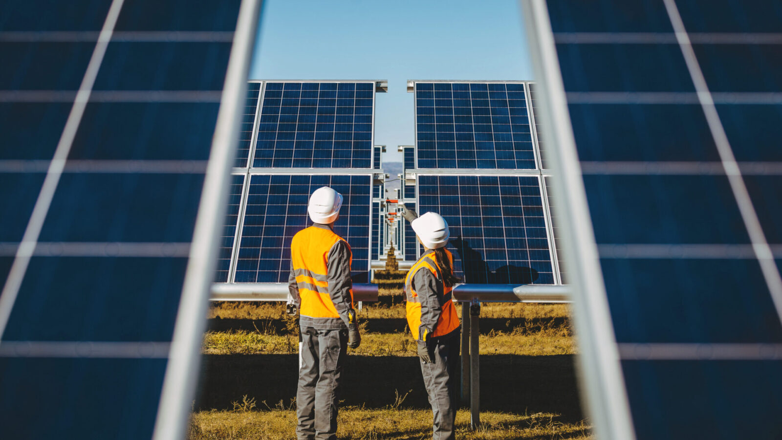 Dos trabajadores con cascos blancos y chalecos de seguridad naranjas se encuentran frente a grandes paneles solares en un campo. El cielo despejado y la luz del sol resaltan la instalación de energía renovable mientras inspeccionan el equipo.