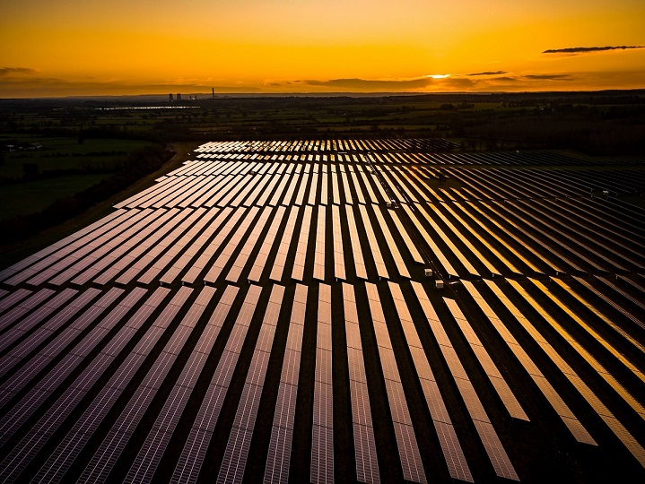 Un vasto campo de paneles solares brilla bajo un cálido y dorado atardecer. Los paneles están dispuestos en filas ordenadas, extendiéndose por el paisaje, con el horizonte y algunas nubes visibles a lo lejos.