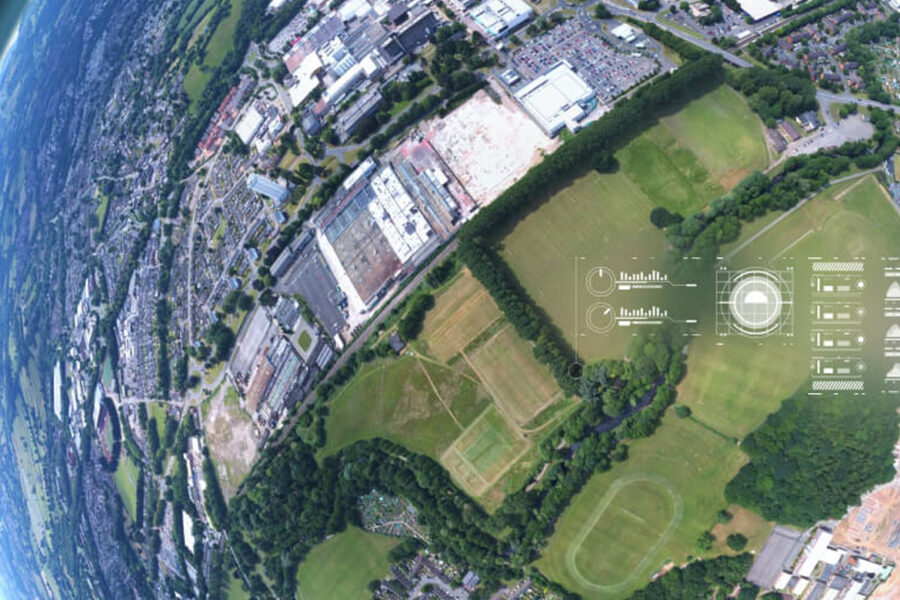 Vista aérea de un paisaje que muestra edificios industriales, campos verdes y zonas residenciales. Una superposición digital presenta gráficos y visualizaciones de datos, que evocan tecnología futurista. El borde de la curvatura terrestre es visible a la izquierda.