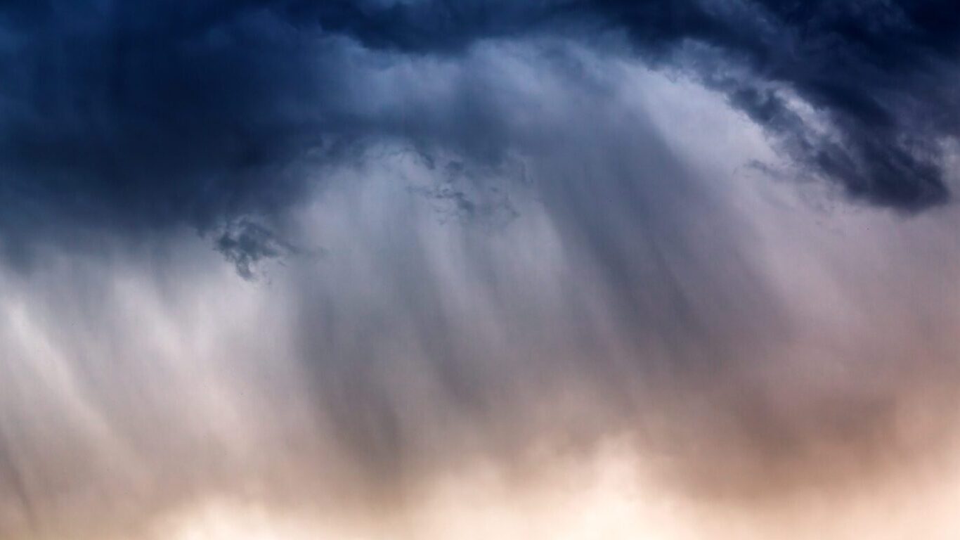 Nubes oscuras de tormenta llenan el cielo con un dramático gradiente que va del azul intenso al gris más claro. La luz del sol ilumina suavemente la parte inferior, creando un efecto brumoso y etéreo que sugiere lluvia intensa y tiempo turbulento.