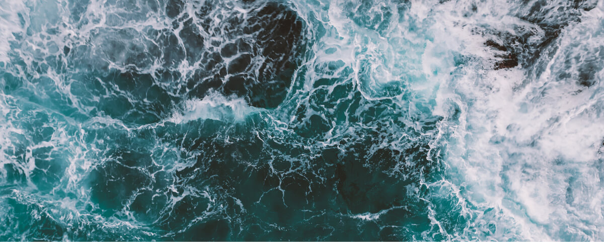 Vista aérea de las turbulentas olas del océano rompiendo y remolineando sobre rocas oscuras, creando un patrón dinámico de espuma blanca y agua azul profundo.