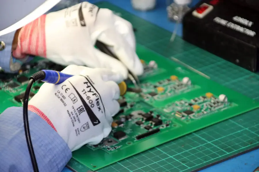 Dos manos enguantadas sueldan componentes electrónicos en una placa de circuito verde. El espacio de trabajo incluye un soldador, una base de corte y varias herramientas. La persona lleva una camisa azul de manga larga.
