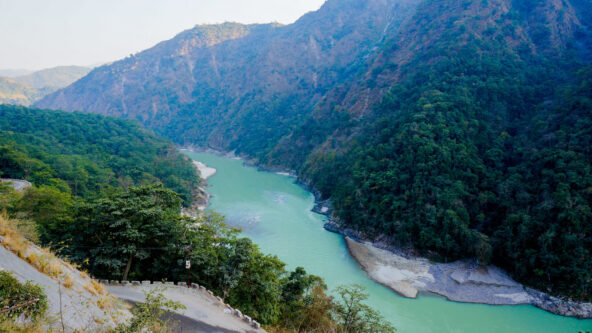 Una vista panorámica de un río serpenteante que fluye entre exuberantes colinas verdes con laderas rocosas. El paisaje está repleto de densos árboles y vegetación, bajo un cielo azul claro. El río crea una atmósfera de paz y serenidad.