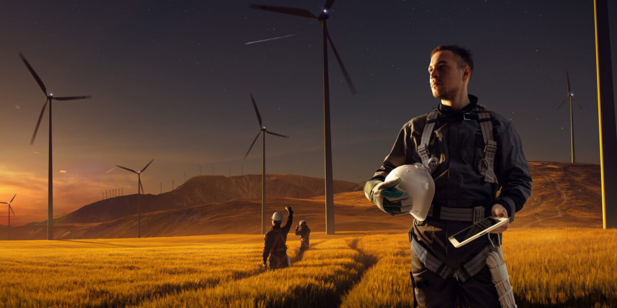 Una persona con uniforme de trabajo sostiene un casco y una tableta, de pie en un campo dorado al atardecer, con aerogeneradores al fondo. Se ven otros trabajadores y una estrella fugaz en el cielo.